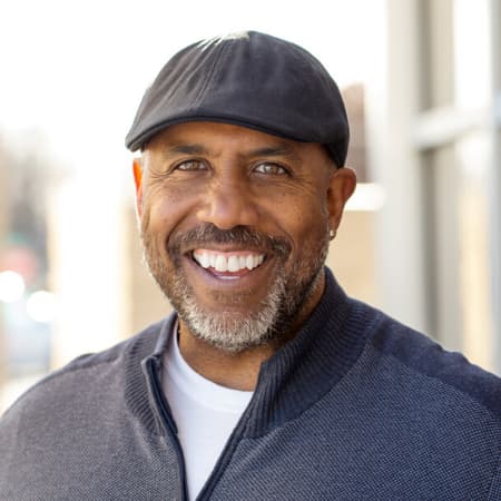 Middled aged black man smiling with gray hat and a gray 3/4 zip jacket on outside a building