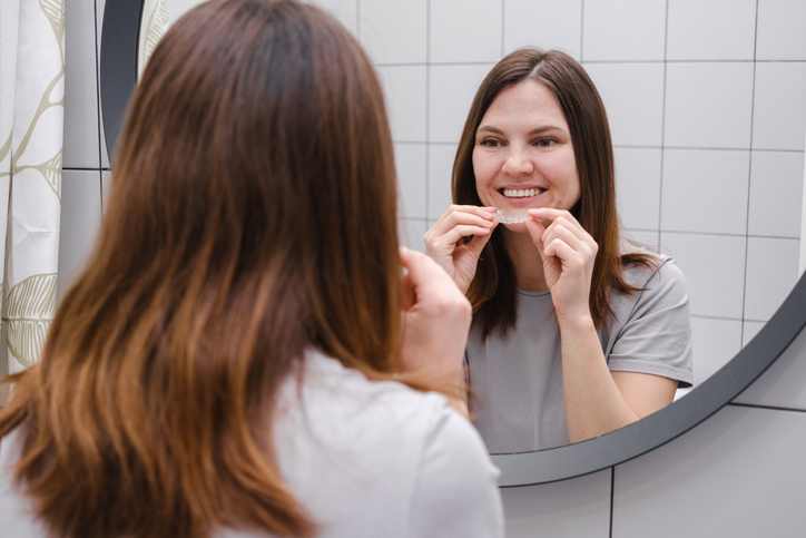 Smiling, dark-haired woman looking in the mirror, holding a clear aligner
